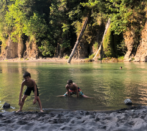 Two boys play in a shallow river, surrounded by trees and rocky cliffs, with one boy swimming in the background.