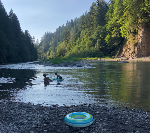 Two children play in a river, surrounded by trees and rocky banks, with a colorful inner tube nearby.