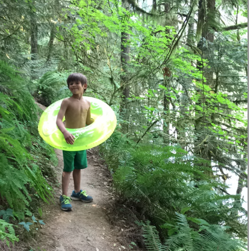 A young boy stands on a forest path, holding a bright yellow inner tube, surrounded by lush green foliage.