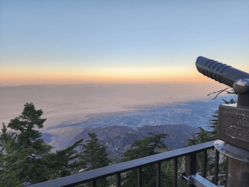 View from a mountain overlook, featuring a telescope and a hazy valley below at sunset. Trees frame the scene.
