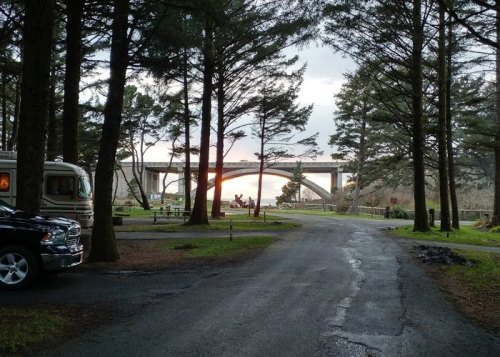 A scenic view of a bridge through tall trees, with a road and parked vehicles in the foreground.