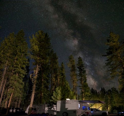 A starry night sky filled with the Milky Way above tall trees and a camper in a forested area.
