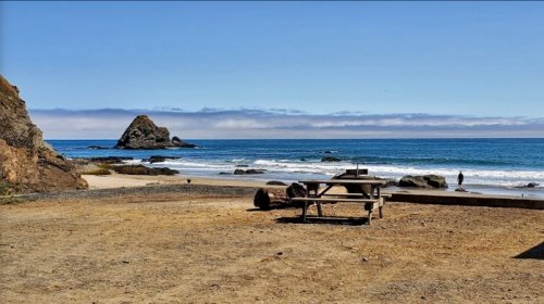 A sandy beach with a picnic table, rocky formations, and gentle waves under a clear blue sky.