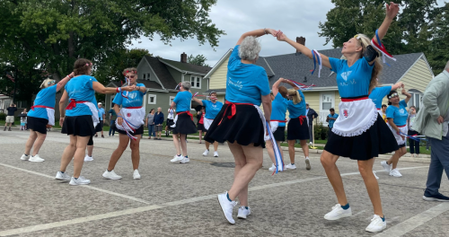 A group of dancers in blue shirts and black skirts perform a lively dance outdoors, celebrating with ribbons and smiles.