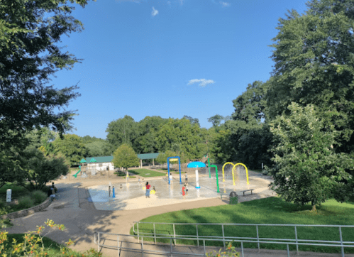 A sunny park scene featuring a splash pad, playground equipment, and trees, with children playing in the water.