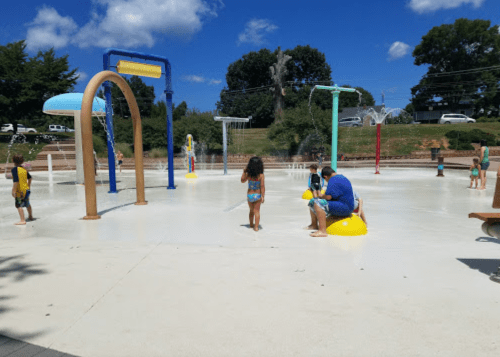 Children playing in a splash pad with water features on a sunny day, surrounded by greenery and blue skies.