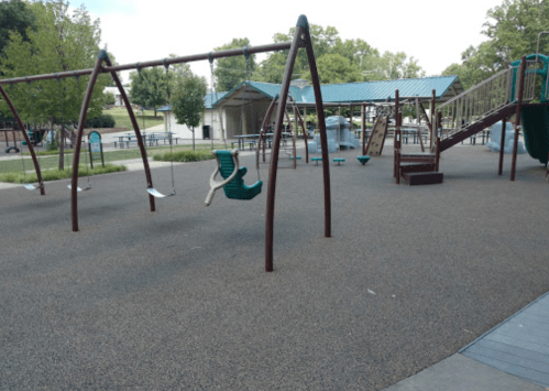 A playground with swings, a slide, and climbing structures, surrounded by trees and a picnic area.