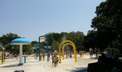 Children play in a splash pad with colorful water features on a sunny day, surrounded by trees and families.