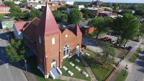 Aerial view of a brick church with a red roof, surrounded by trees and a parking lot in an urban area.