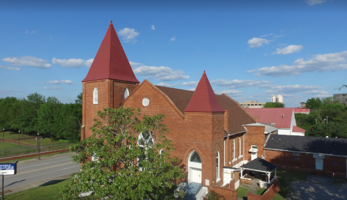 A red-brick church with two pointed towers, surrounded by trees and a clear blue sky with scattered clouds.