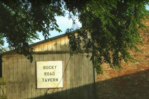Sign for "Rocky Road Tavern" on a rustic wooden building, framed by green leaves and soft sunlight.