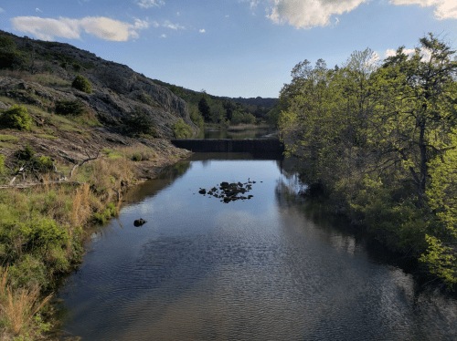 A serene river scene with rocky banks, trees, and a calm water surface reflecting the sky.