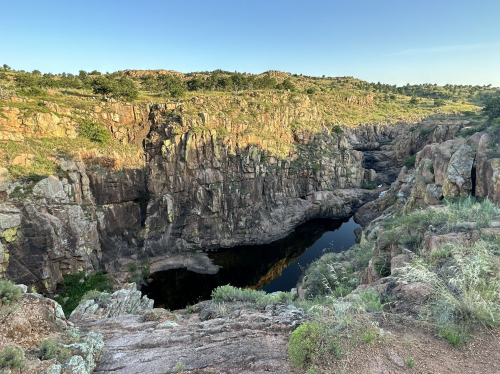 A rocky canyon with steep cliffs and a dark pool of water at the bottom, surrounded by greenery and sunlight.