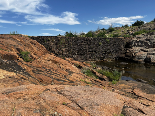 A rocky landscape with a stone wall and a calm water body under a blue sky with scattered clouds.