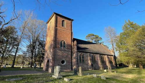 Historic brick church surrounded by trees and gravestones under a clear blue sky.