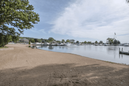 A sandy beach area by a calm lake, with boats docked and trees lining the shore under a cloudy sky.