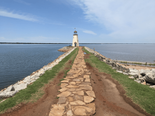 A stone pathway leads to a lighthouse on a rocky pier, surrounded by calm water and a clear blue sky.
