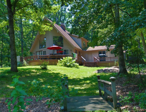 A spacious house surrounded by trees, featuring a deck with an umbrella and a small wooden bridge in the foreground.