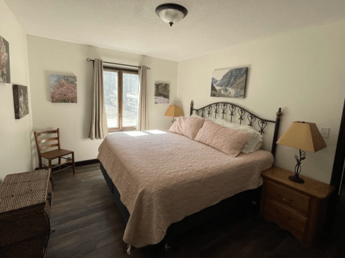 Cozy bedroom with a bed, nightstand, lamp, chair, and nature-themed wall art near a window.