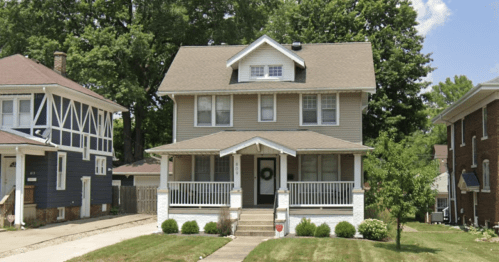 A two-story house with a front porch, surrounded by greenery and a well-maintained lawn on a sunny day.