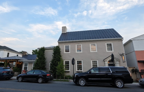 A two-story gray house with a metal roof, flanked by trees, and parked cars on a street under a blue sky.