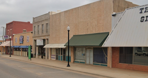 A quiet street scene featuring several storefronts with awnings and a lamppost, under a cloudy sky.