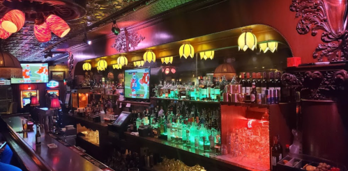 A vibrant bar scene with colorful lighting, shelves of liquor, and screens displaying sports. Cozy atmosphere.