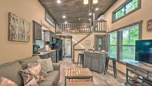 Cozy interior of a modern cabin with a kitchen, living area, and loft, featuring large windows and wooden accents.