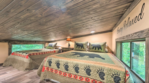 Cozy loft bedroom with two beds, wooden ceiling, and bear-themed decor, surrounded by large windows.
