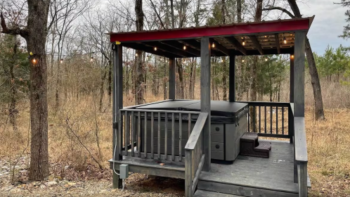 A wooden gazebo with a hot tub, surrounded by trees and a grassy area, under a cloudy sky.