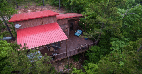 Aerial view of a rustic wooden cabin with a red metal roof, surrounded by lush green trees and a wooden deck.