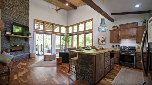 A modern kitchen with a stone island, wooden beams, large windows, and a cozy seating area.