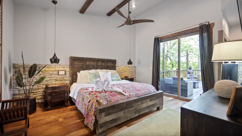 Cozy bedroom with a wooden bed, colorful bedding, stone accent wall, and large windows overlooking a green outdoor space.