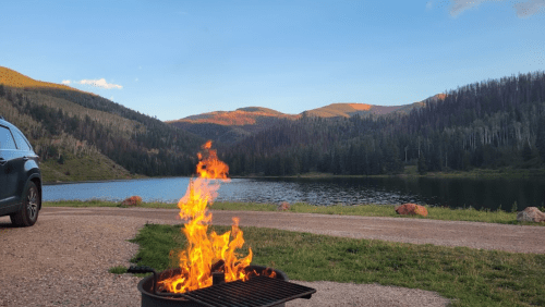 A campfire burns in a grill by a serene lake, surrounded by mountains and trees under a clear blue sky.