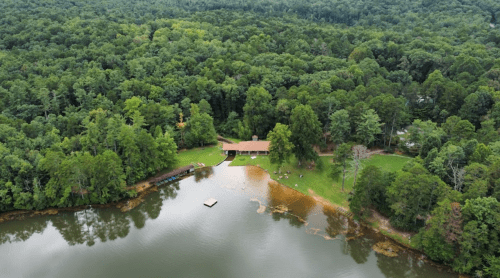 Aerial view of a house by a lake, surrounded by lush green trees and a small dock on the water.