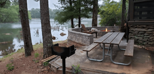 A lakeside campsite with a picnic table, fire pit, and stone seating area surrounded by trees and water.