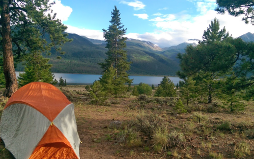 A tent set up by a serene lake, surrounded by trees and mountains under a partly cloudy sky.