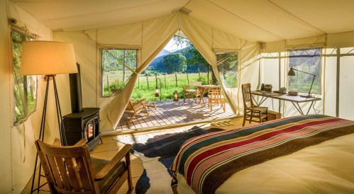 Cozy glamping tent interior with a bed, desk, and outdoor seating, surrounded by nature and sunlight.