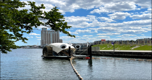 A unique boat with large round features docked by a waterfront, under a blue sky with fluffy clouds and a city skyline in the background.