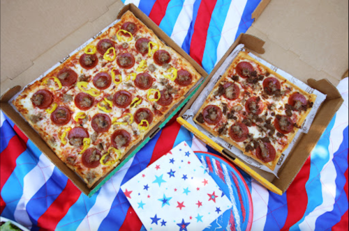 Two pizza boxes on a colorful tablecloth: one large pepperoni pizza and one smaller pizza with sausage.