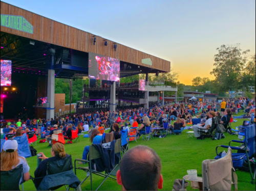 A large crowd enjoys a concert at an outdoor venue, with a stage and large screens, under a colorful sunset.