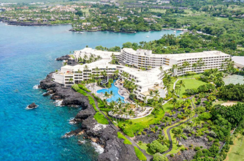 Aerial view of a coastal resort with palm trees, pools, and rocky shoreline, surrounded by lush greenery and blue water.