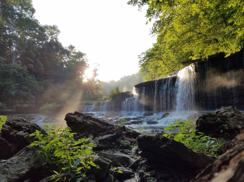 A serene waterfall surrounded by lush greenery, with sunlight filtering through the trees and mist rising from the water.