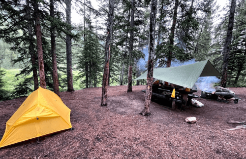 A yellow tent and a cooking area under a tarp, surrounded by tall trees in a forested campsite. Smoke rises from the fire.