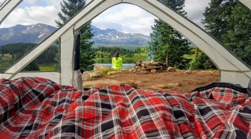 A view from inside a tent, showing a person meditating by a lake surrounded by mountains and trees.