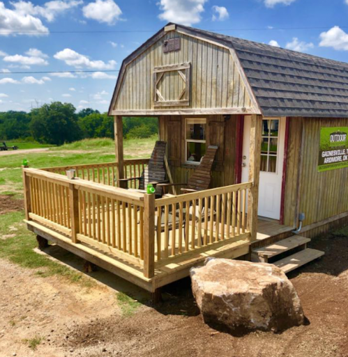 A rustic wooden cabin with a porch, surrounded by grass and blue skies, featuring a sign for an outdoor store.