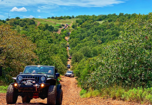 A line of off-road vehicles driving on a dirt trail through a lush, green landscape under a blue sky.