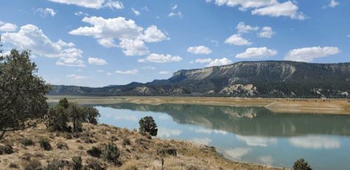 A serene landscape featuring a calm lake reflecting clouds and mountains, surrounded by sparse vegetation and trees.
