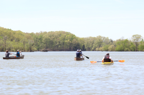 Four people in kayaks and canoes paddle across a calm lake surrounded by lush green trees.