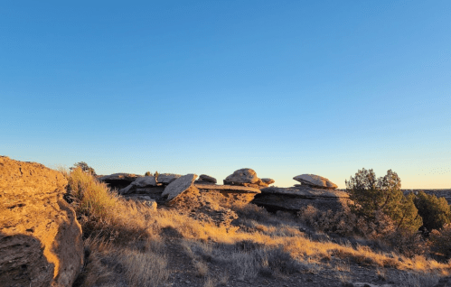 A rocky landscape with large boulders under a clear blue sky, surrounded by sparse vegetation.